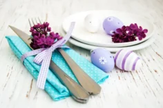 A imagem apresenta uma elegante mesa posta de Páscoa. Talheres dourados estão sobrepostos a um guardanapo azul piscina com bolinhas brancas, envoltos a um laço branco e lilás. Além disso, a decoração conta com ovos coloridos em tom lilás, pratos brancos e flores roxas.